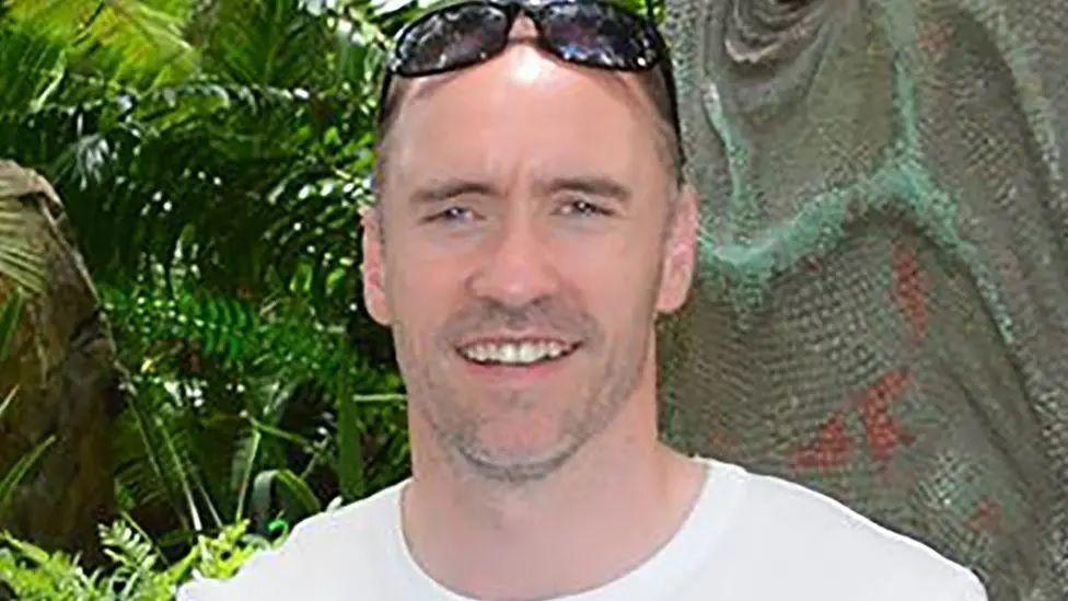 Paul Doyle has short dark greying hair with sunglasses on his head and is wearing a white T-shirt.