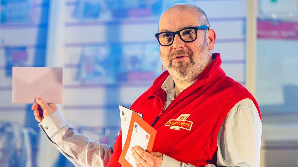 A cast member in a red Post Office uniform holding up several letters