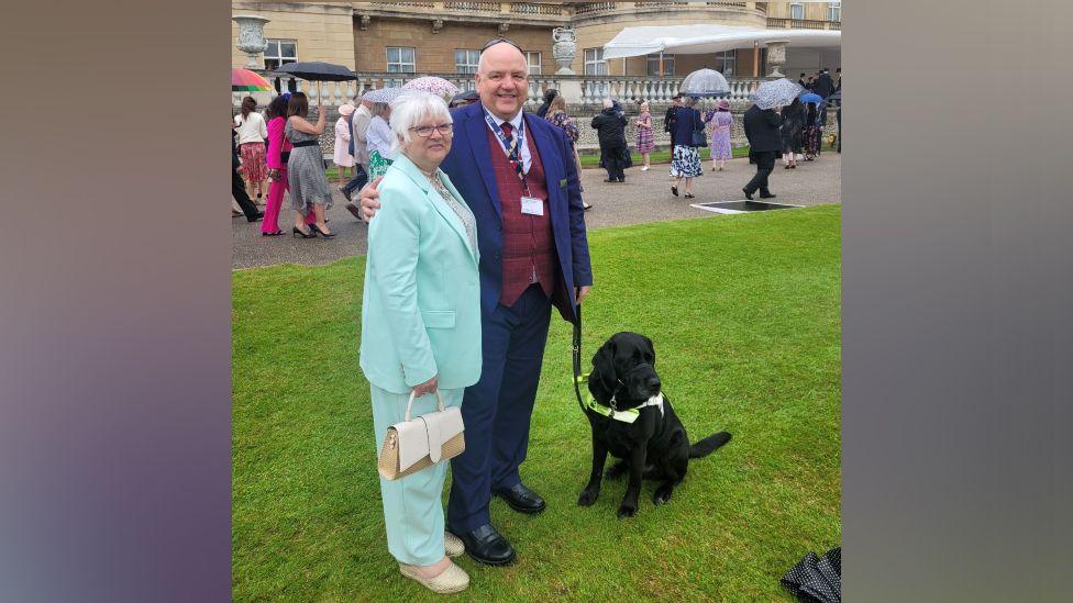 John Hardy with his arm around his wife's shoulders. They are both in suits standing in the garden of Buckingham Palace. John is holding Sidney, a black Labrador, on a lead.