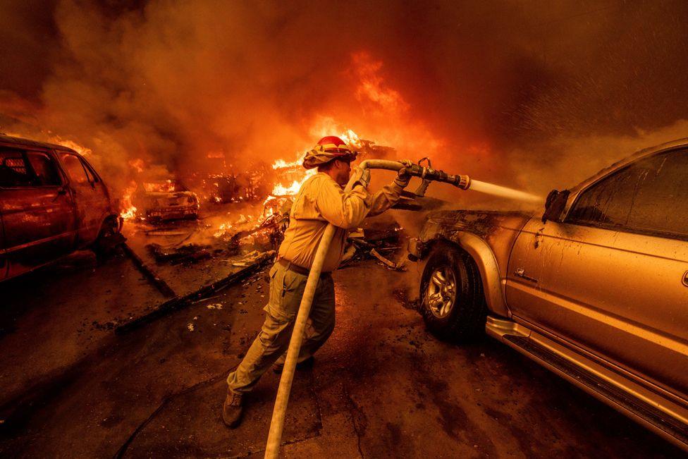 Flames can be seen in the background as a firefighter lifts a water hose to spray a burning vehicle