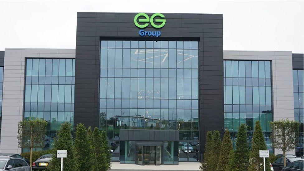 The EG group headquarters, a large building with a rectangular grey frame and lots of glass windows. In the middle is a taller black framed structure, also with many windows and the EG group logo in green and blue at the top
