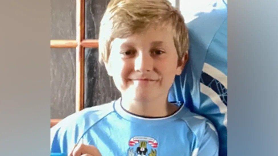 A young boy wearing a Coventry City shirt. He has short blonde hair, parted at the side, and the shirt is light blue with a crest on the front. Part of a glass-panelled wooden door is visible behind him