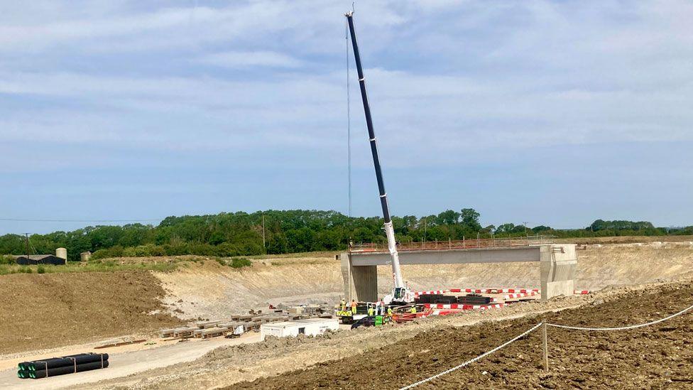 The Black Cat roadworks. The road is currently a cutting in the ground, showing yellow chalky soil. At the bottom of the cutting is a flat white service, various concrete rafters, people in high-vis working with a road work machine with a crane at its front. Just behind them is a section of a concrete bridge. Beyond are trees and blue skies.