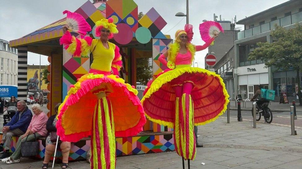 Two people on stilts and wearing neon yellow and pink costumes. 