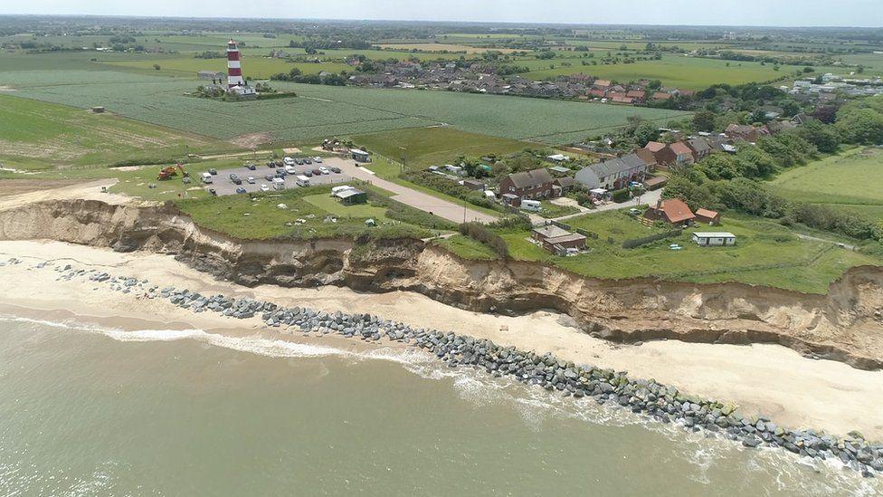 A large stretch of the coastline at Happisburgh. The sea laps against a long row of rocks. Above the sandy cliffs, fields and small villages stretch into the distance. Happisburgh lighthouse can also be seen