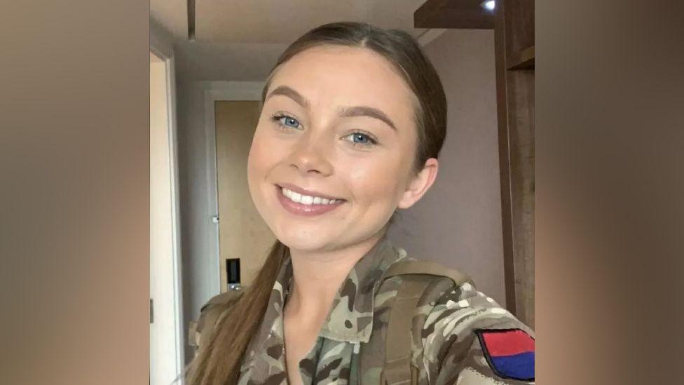 A selfie of Jaysley Beck wearing her military uniform, with her long brown hair slicked back into a low ponytail. She has blue eyes and is smiling at the camera.