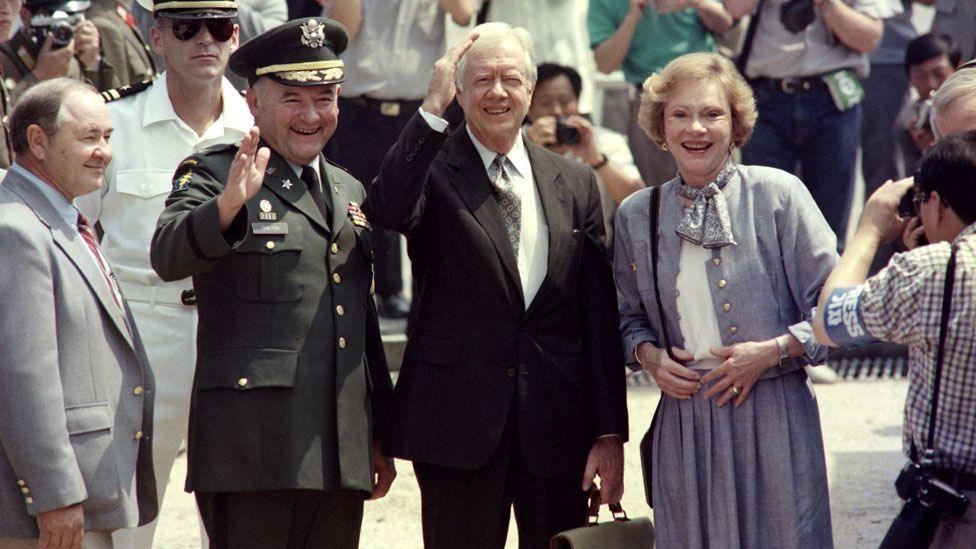 Former U.S. President Jimmy Carter and his wife Rosalynn wave to journalists, surrounded by a crowd. He wears a dark suit, and she wears blue skirt, jacket and scarf