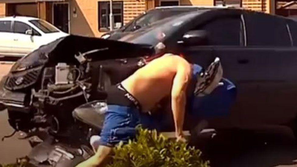 A black car which has driven into a man on a scooter. The vehicle's bonnet has shattered on contact. The man, who is topless, is bending over the side of his scooter in response to being struck.