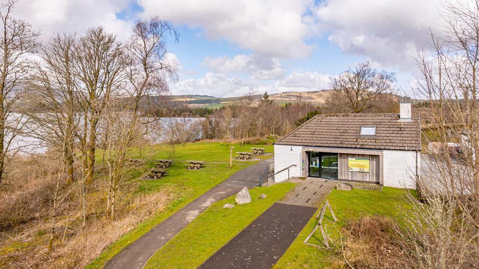 A small white building at the end of a curved path in the middle of wild countryside and next to a loch