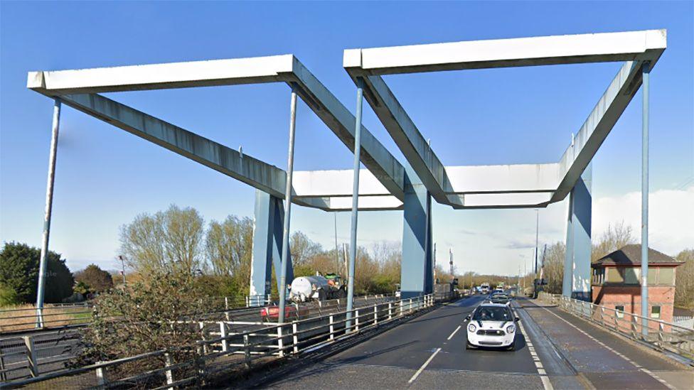 A white car drives across the Ennerdale Bridge which consists two blue and white metal frames with two lanes of traffic either side