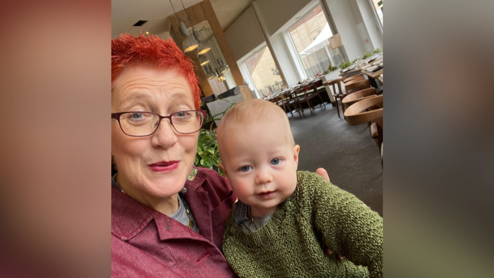 Self taken portrait photo of Fiona Carey with her young grandson. She has blue eyes and is looking at the camera. Her grandson, who also has blue eyes, is sat on her knee wearing a khaki-green jumper. They appear to be sat in a canteen or restaurant, as there are tables and chairs in the background. 