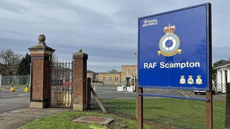 A view from the gates of RAF Scampton with a sign in the foreground, with RAF buildings  behind