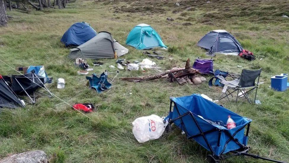 A green open space near trees littered with plastic shopping bags and abandoned camping equipment, including tents and canvas folding chairs.