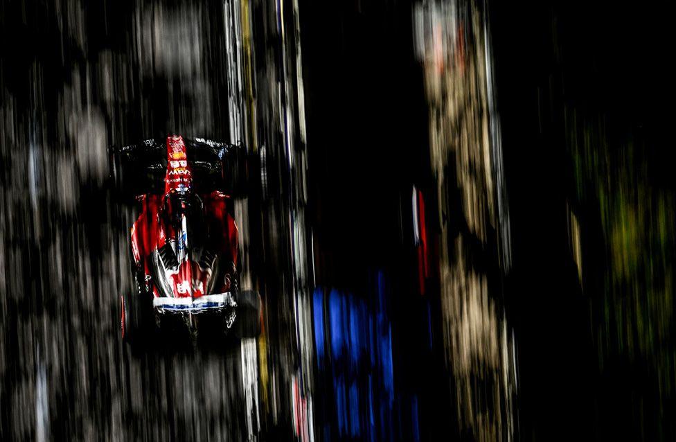 Charles Leclerc drives a red Scuderia Ferrari Formula 1 car during practice for the Singapore Grand Prix at the Marina Bay Street Circuit. The image, taken from above, shows the car racing at high speed with motion blur emphasizing its aerodynamic shape on the narrow track.
