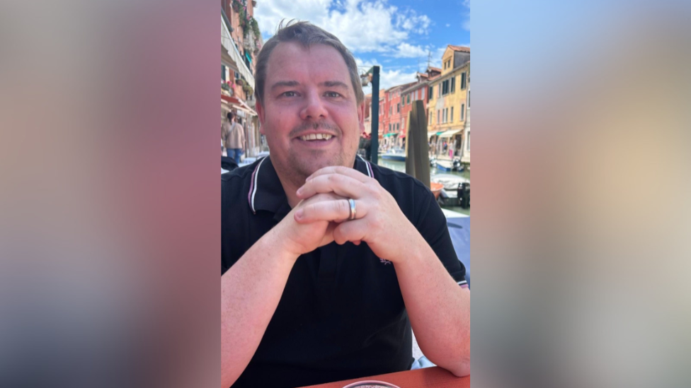 A man is sat at a table smiling into the camera with his hands clasped together in front of his chest. He appears to be in a city on a sunny day.
