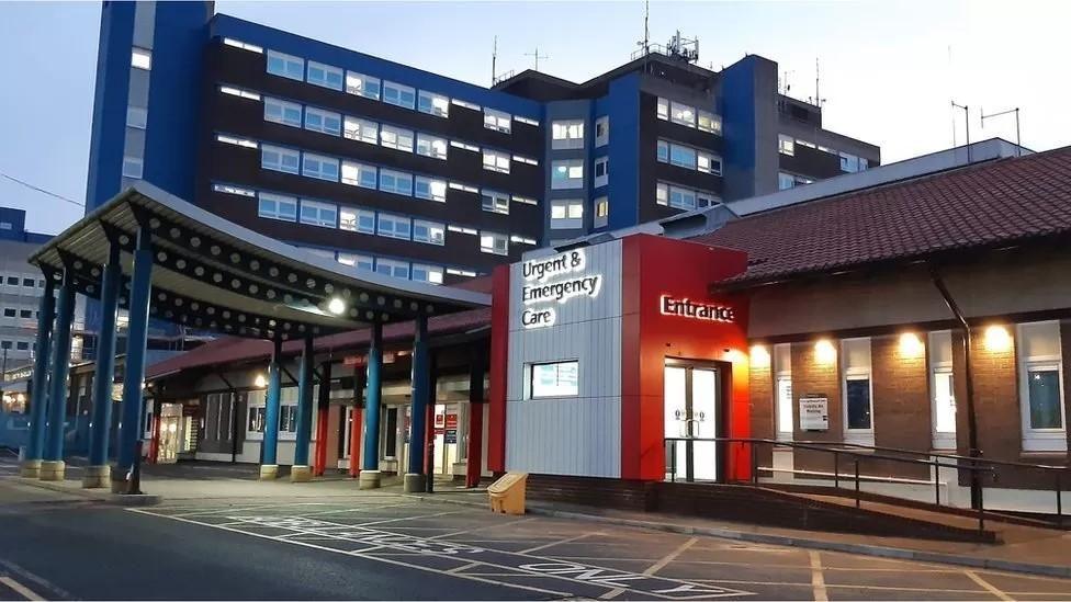 A well lit hospital entrance with a larger building in the background and a canopy held up by blue pillars over the door 