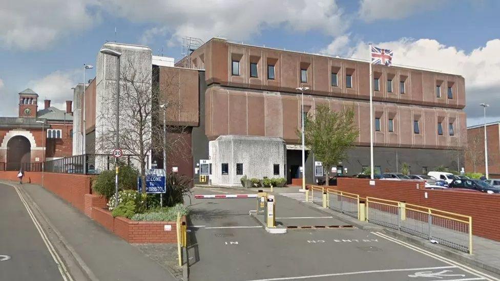 A Google Streetview image of HMP Bristol, showing the vehicle access gate and the front of the prison. The structure is a large, block-like building, with a Union Jack flying in the car park area. 