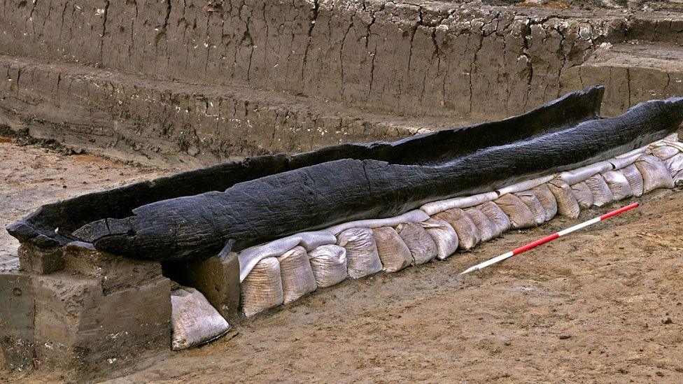 A blackened log boat at Must Farm quarry after excavation. It is hollowed out in the centre and is resting on a bed of sand bags. Behind it can be seen a trench wall. 