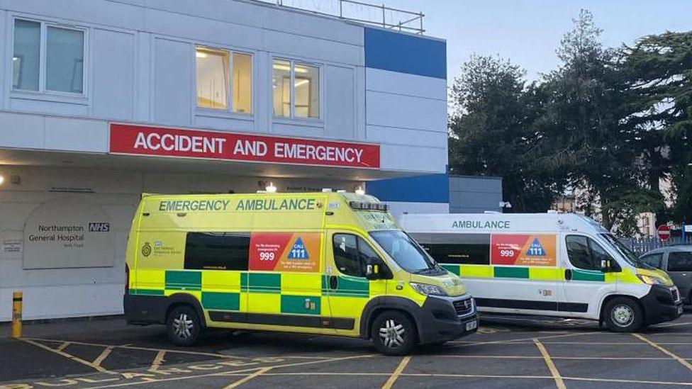 A yellow and green ambulance and a white ambulance parked outside a white coloured Accident and Emergency building at Northampton General Hospital.