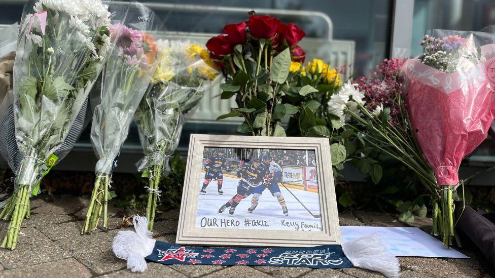 Flowers, a scarf and a picture on a pavement propped up against a window. The framed picture is of ice hockey players with the message "our hero" written on it.