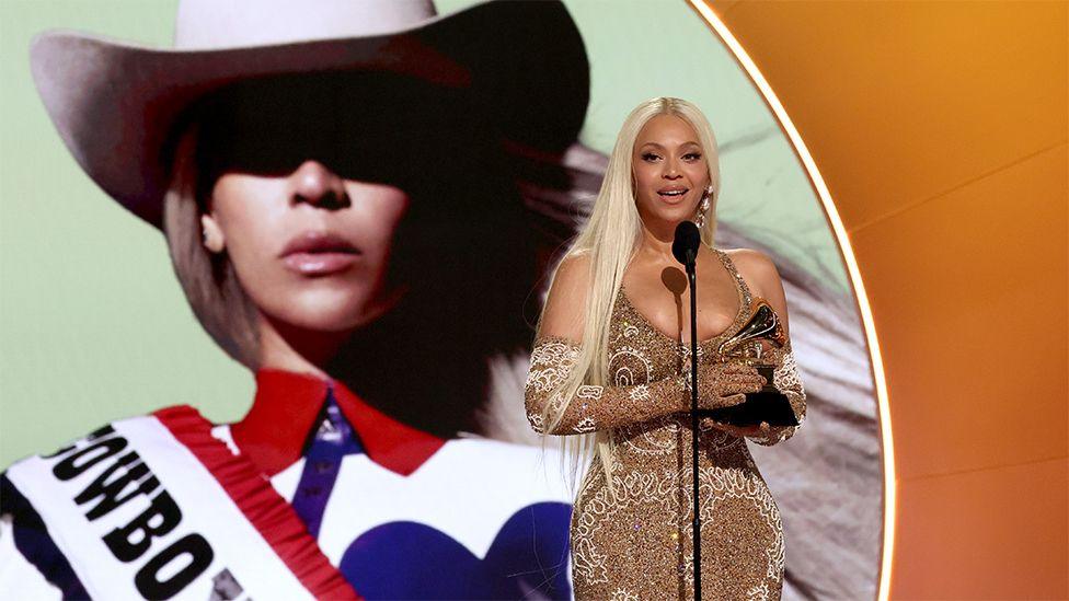 Beyonce accepting a grammy award, speaking into a black microphone while wearing a sparkling outfit. Behind her is a picture of herself with a "Cowboy Carter" poster.