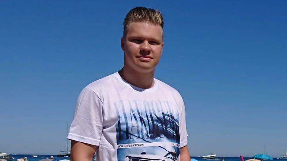 A young man with slicked-back blond hair in a white T-Shirt with a blue logo on it on a lovely sunny day on the beach