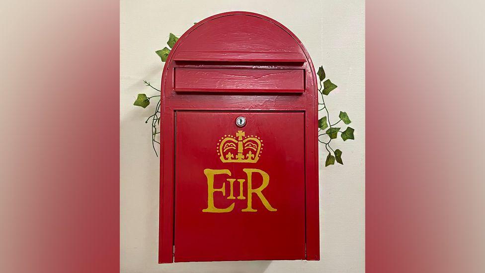 A wooden post box is painted red and fixed to a white wall. It has a gold crest and the initials ER on the front, indicating Queen Elizabeth II. A sprig of ivy is placed on top as decoration.