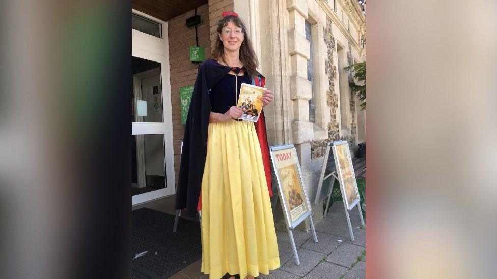 Ms Moseley pictured outside the theatre dressed as Snow White while holding a theatre programme. She has long brown hair and is wearing a yellow floor-length skirt with a navy blue top and navy blue cape.