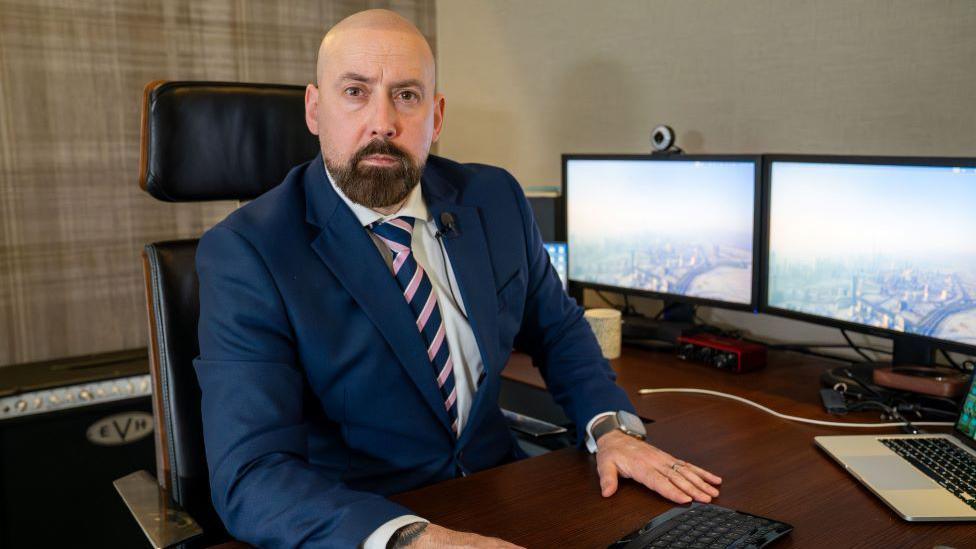 David Kennedy in a suit looking at camera. He is sitting in front of computer and screens.