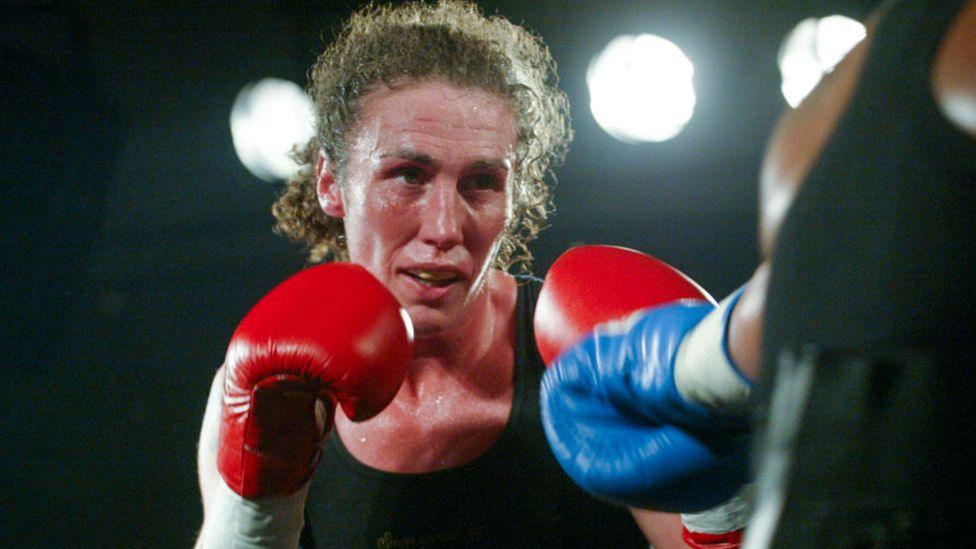 Couch boxing in a black vest with her brown curly hair tied back and red boxing gloves held up to her chin