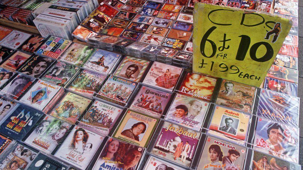 A stall displaying a large collection of CDs and casette tapes laid out face up, displaying their covers based on Bollywood film posters. A tattered neon yellow sign stands in the middle of the collection with the words "CDs, 6 for £10, £1.99 each" written in large black letters.