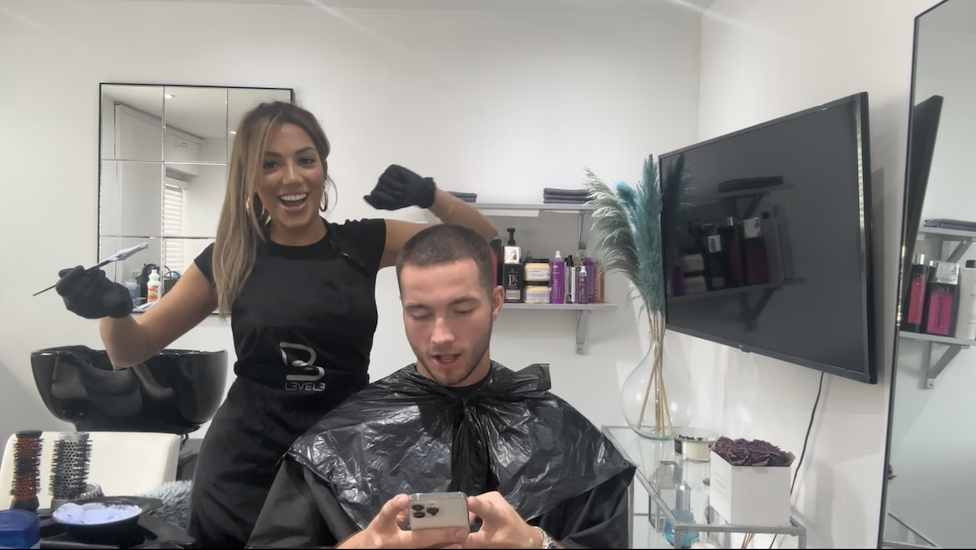 Tanyel wears a black outfit and gloves, whilst Ron is also wearing black whilst sitting down in a chair in the salon.