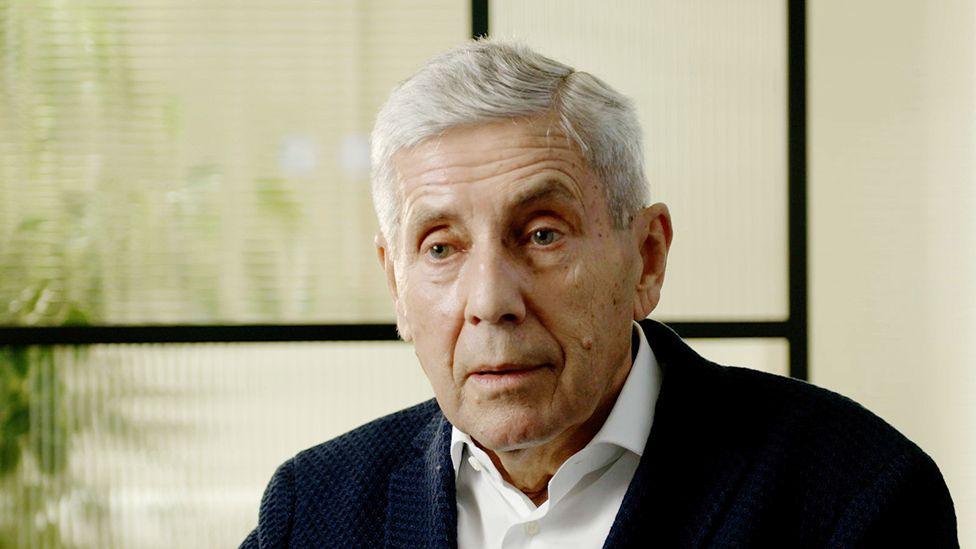 Lord Rose, a man with silver hair swept in a side-parting, wearing a crisp white shirt and a dark jacket with a subtle woven pattern. He is sitting in front of a black-framed screen with ridged glass, which distorts the view of a pot plant behind it.