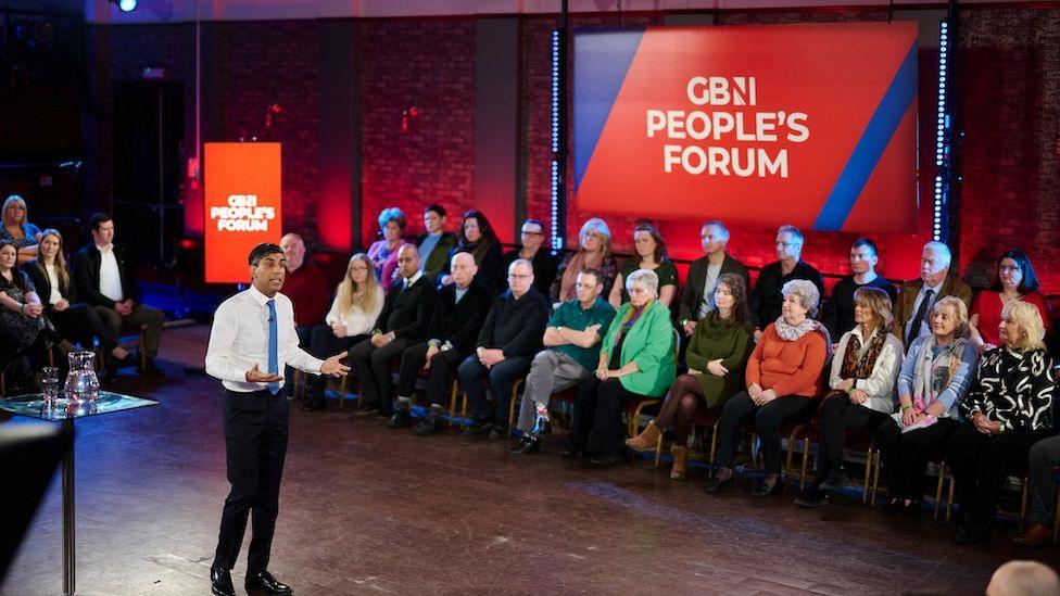 Former Prime Minister Rishi Sunak, wearing a suit with a white shirt and blue tie, appears in front of an audience of people and a sign which reads "GB People's Forum".