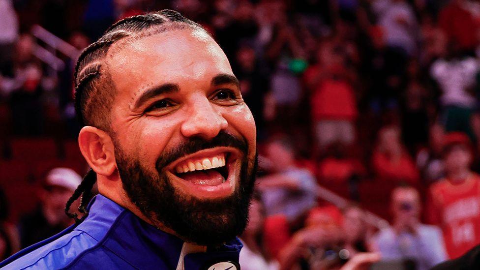 Drake, smiling, wearing a blue top, with fans behind him at a basketball game out of focus