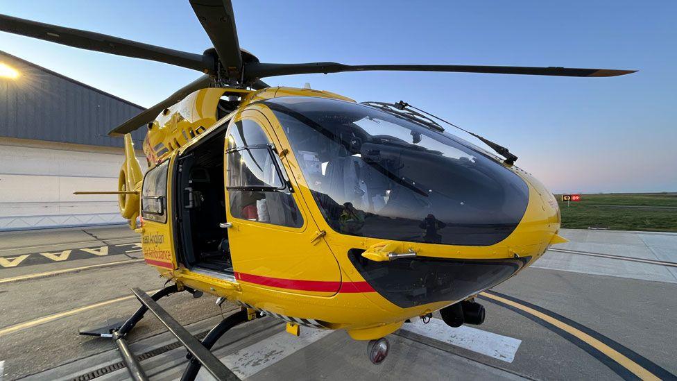 An East Anglian Air Ambulance helicopter in its bright yellow livery. It is resting on a runway with a hangar behind it.