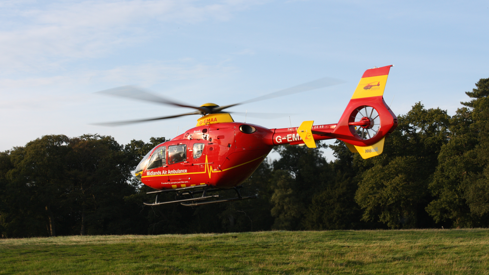 A Midlands Air Ambulance helicopter in flight. The helicopter is red and yellow and is pictured a short distance above the ground shortly after take off on a sunny day with trees in the background.