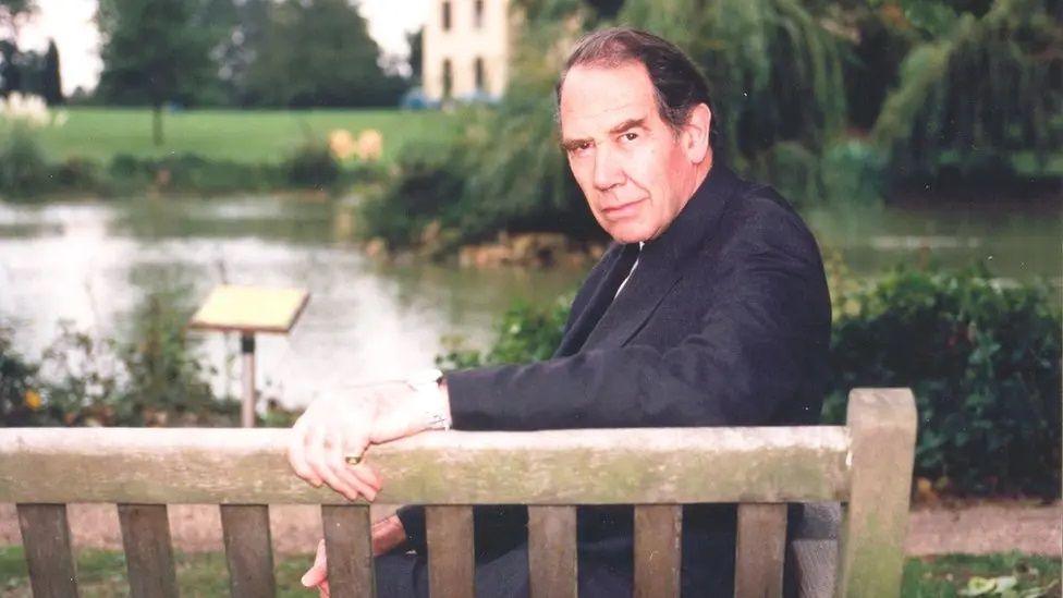 Sir Terence English sitting on a bench, looking back towards the camera. His left hand is resting on the back of the wooden bench. He is wearing a dark jacket. There is a lake and a house in the background.