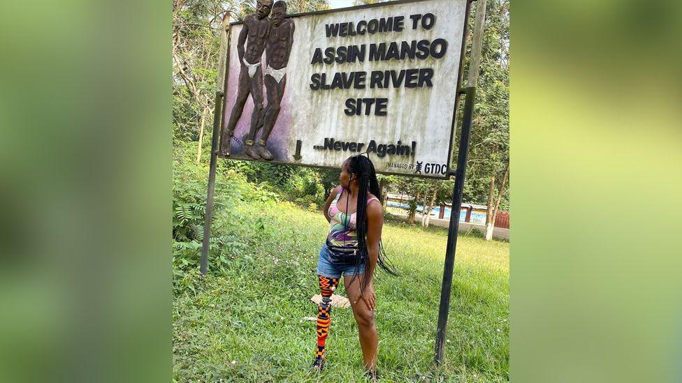 Abena Christine Jon'el  standing on some grass in front of a sign that that says "welcome to Assin Manso Slave River site". She is turning towards the sign and her kente covered prosthetic leg can be seen.