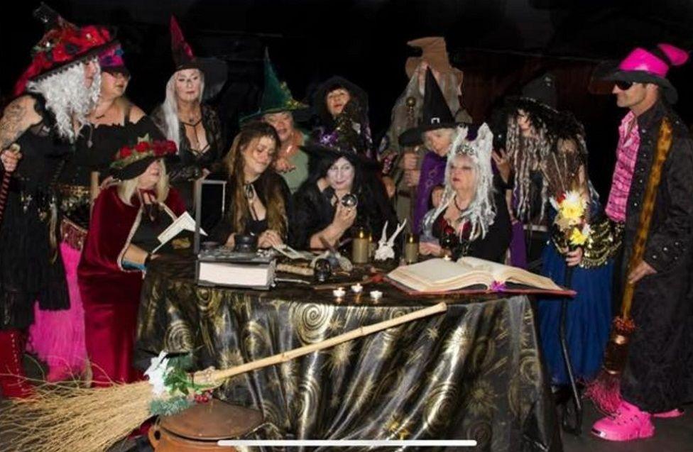 Members of the Boston Dancing Witches wearing their outfits and sat round a table with their spell books and a broom. They are wearing different coloured cloaks and hats.