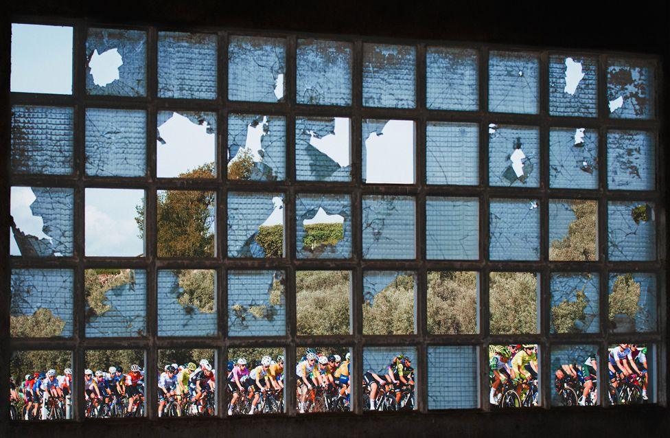 A large group of cyclists in colourful jerseys seen through a shattered glass window. The broken panes frame the riders against a backdrop of trees and sky.