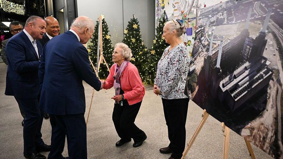 An elderly woman curtseys as she shakes King Charles's hand, as others look on