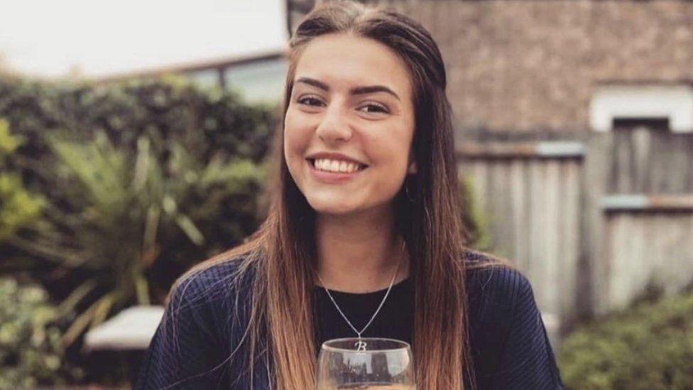 A young woman with long brown hair smiles at the camera while holding a wine glass. She is wearing a blue long sleeved top with a necklace around her neck. 