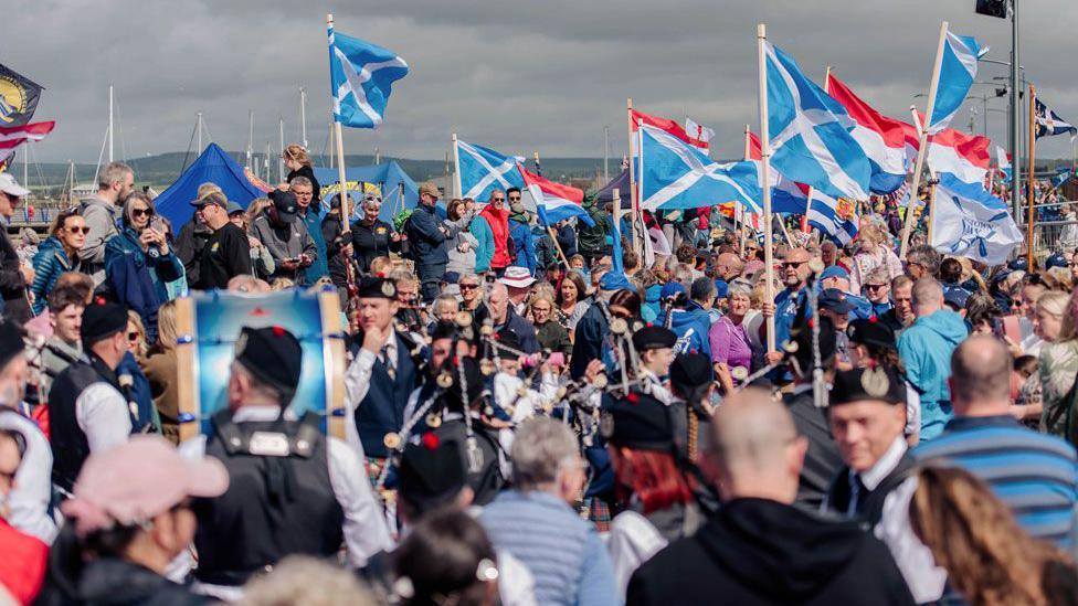 A huge crowd scene with flags, pipers, drummers and spectators