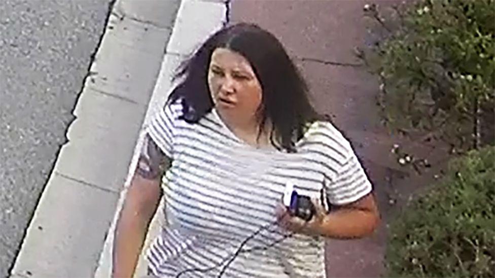 A woman with dark hair and a serious expression. She wears a striped black and white shirt. On her right arm there appears to be a tattoo of a snake and she is carrying several items in her left hand.