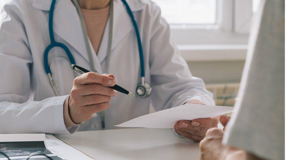 An anonymous doctor wearing a white doctor's coat with a stethoscope around their neck while they sit at a desk with a pen in one hand and a piece of paper in the other.