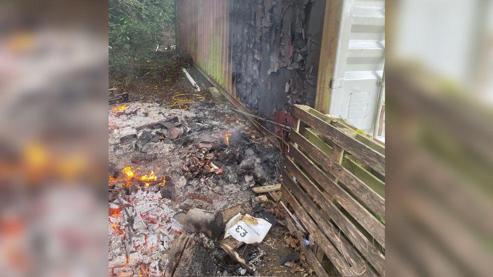 The exterior of a storage container. The paint on the metal corrugated walls has been blackened and is peeling off. On the ground there is a pile of grey ashes still on fire and a wooden pallet leading up against the container.