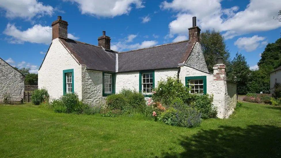 A whitewashed stone farmhouse with green window frames and a slate roof beneath a blue sky with a few white clouds