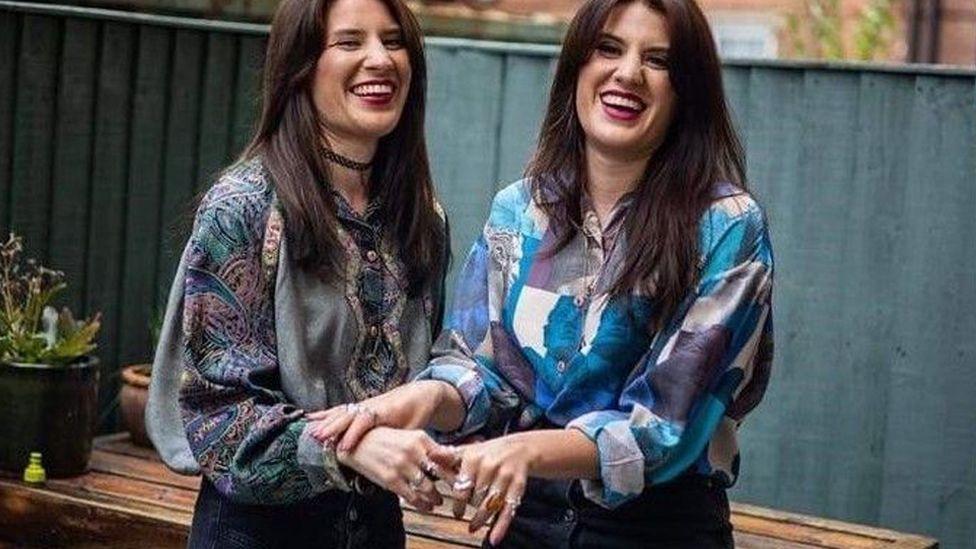 Georgia and Melissa with a garden fence behind them. They are both wearing shirts and denim skirts.