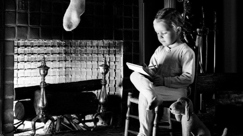 A black and white image of a girl of about five or six years old, sitting on a chair beside a fireplace. She is resting her right leg across her left knee and has a notepad resting on the leg on which she is writing. Above the fireplace is hanging the bottom half of a stocking